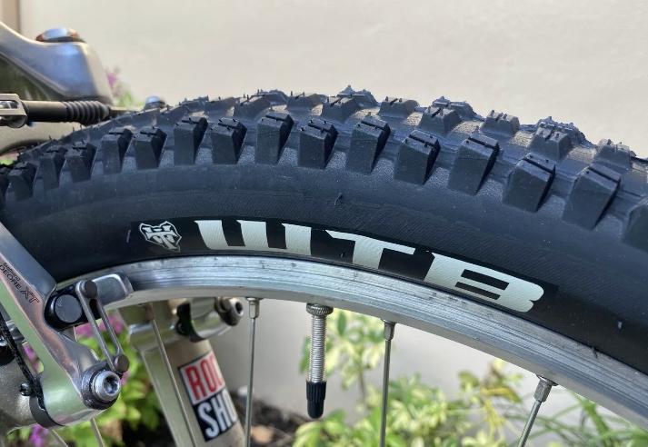 Specialized Specialized Stumpjumper A1 FS Mountain Bike: Close-up image of a mountain bike tire featuring a prominent "WTB" logo, showcasing the rugged tread pattern designed for traction. The tire is mounted on a bike wheel with visible components, including a RockShox shock absorber in the background.