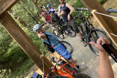 A group of cyclists posing on a wooden bridge in a wooded area, with several mountain bikes lined up beside them. The foreground features a person with a helmet taking a selfie and smiling, while others in the background are giving gestures of excitement. The scene captures a fun outdoor biking adventure surrounded by greenery. Cub Creek Trails mountain bike trail.