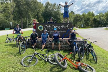 A group of seven mountain bikers poses in front of the Cub Creek Park sign. They are smiling and wearing biking gear, with colorful helmets and jerseys. Some bikes are leaned against the stone marker, while others lie on the grass. The park features trees and a sunny sky in the background, creating a vibrant outdoor atmosphere. Cub Creek Trails mountain bike trail.