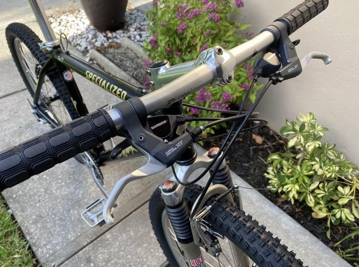 Specialized Specialized Stumpjumper A1 FS Mountain Bike: Close-up view of a mountain bike's handlebars, featuring black textured grips, Shimano brake and gear levers, and a silver handlebar stem. The bike is positioned on a paved surface with green plants and flowers in the background.