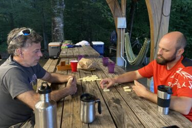 Two men are sitting at a wooden table covered with dominoes. The man on the left has curly hair and is wearing a gray t-shirt, while the man on the right has a bald head and a beard and is wearing a red t-shirt. Both are focused on the game. Various drink containers and a bag of snacks are on the table, and a hammock can be seen in the background surrounded by trees. The setting appears to be outdoors in the evening. Cub Creek Trails mountain bike trail.