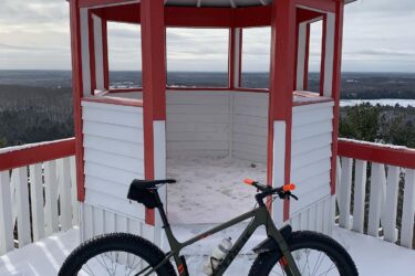A fat tire bike parked beside a snow-covered lookout tower with red and white trim, surrounded by a winter landscape featuring snow and cloudy skies in the background. Elliot Lake Mountain Bike Trails mountain bike trail.