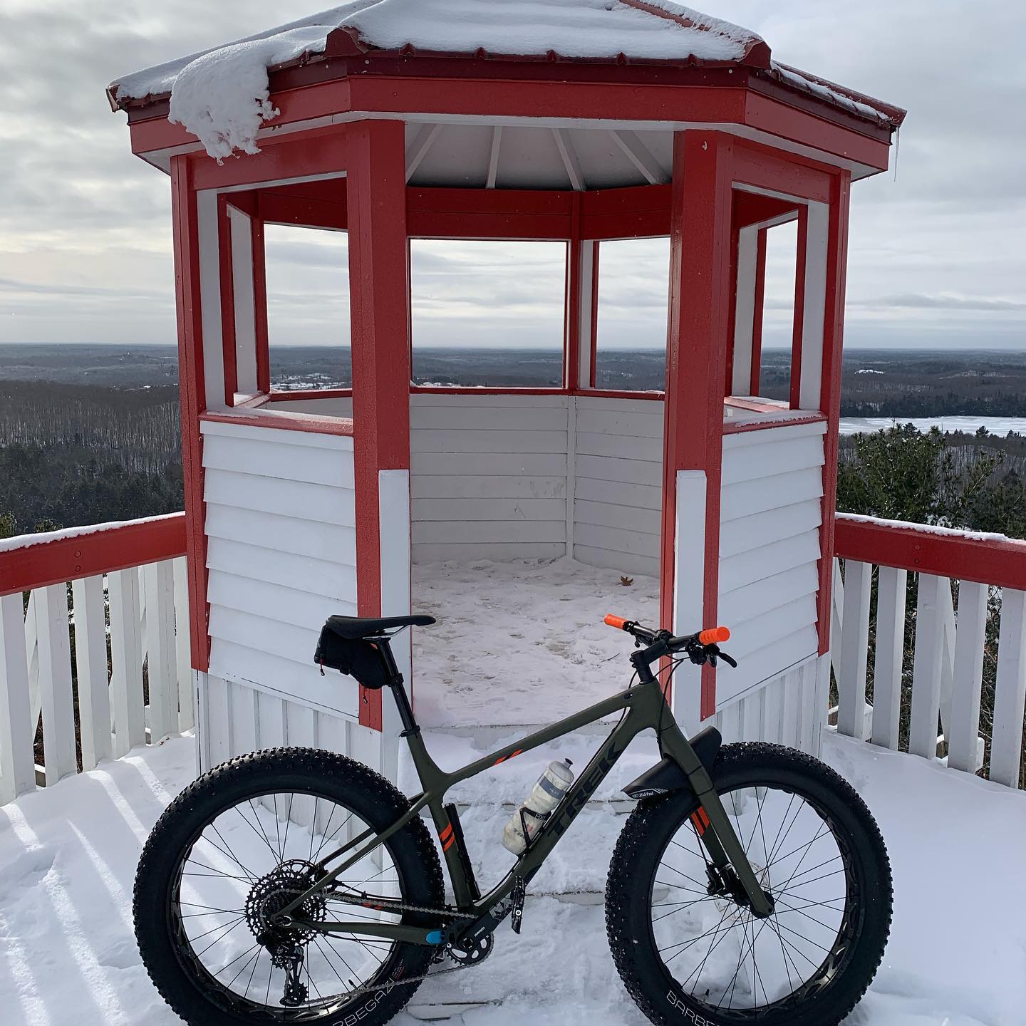 A fat tire bike parked beside a snow-covered lookout tower with red and white trim, surrounded by a winter landscape featuring snow and cloudy skies in the background. Elliot Lake Mountain Bike Trails mountain bike trail.