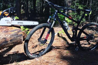 A mountain bike parked next to a chainsaw on a fallen log, set in a forested area with sunlight filtering through the trees. Elliot Lake Mountain Bike Trails mountain bike trail.