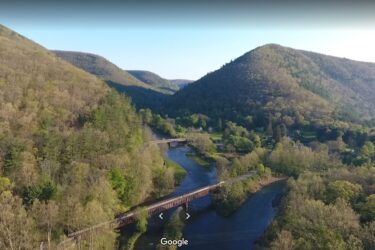 Aerial view of a serene landscape featuring rolling green hills and a winding river, with two bridges crossing the water. The scene is bathed in soft sunlight, highlighting the lush vegetation and tranquil atmosphere of the area. REYNOLDS SPRING NATURAL AREA - LARIAT mountain bike trail.