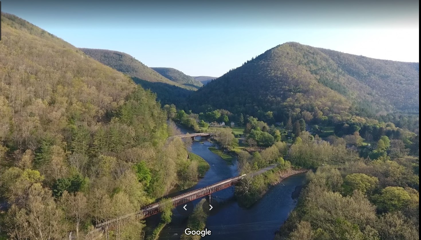 Aerial view of a serene landscape featuring rolling green hills and a winding river, with two bridges crossing the water. The scene is bathed in soft sunlight, highlighting the lush vegetation and tranquil atmosphere of the area. REYNOLDS SPRING NATURAL AREA - LARIAT mountain bike trail.