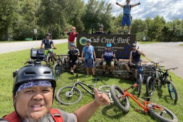 A group of eight mountain bikers posing together at Club Creek Park, smiling and showing thumbs-up gestures. Several bicycles are parked nearby on the grass, while one member is playfully standing on the sign of the park. The background features trees and a clear sky, indicating a sunny day outdoors. Cub Creek Trails mountain bike trail.