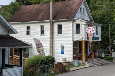 A white two-story building with a brown roof, featuring a large ice cream cone sign out front. The building is situated near a road with greenery and a hillside in the background. There are steps leading up to the entrance, and a small fenced area with landscaping in the foreground.  REYNOLDS SPRING NATURAL AREA - LARIAT mountain bike trail.
