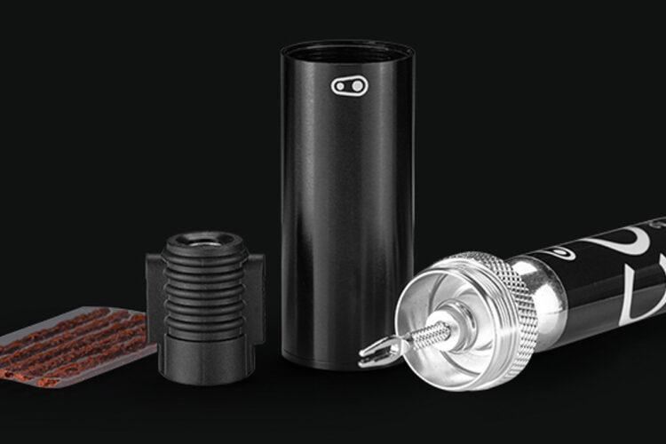 A collection of smoking accessories displayed on a dark background, including a black cylindrical case, a black connector piece, a small metal tool, and a glass tube with a black label.