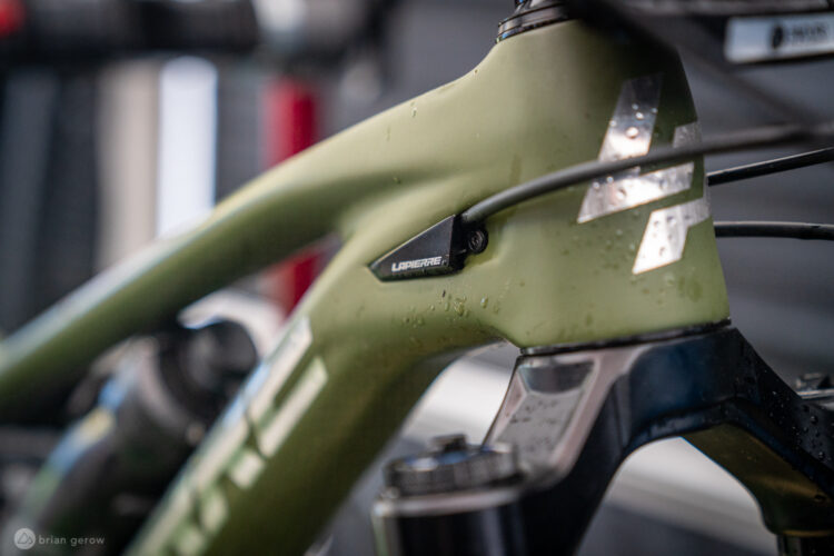 Close-up of a bicycle frame with a green finish, featuring a Lapierre branding badge. The frame shows water droplets, suggesting recent exposure to moisture. Blurred background indicates a workshop or garage setting.