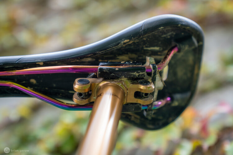 Close-up view of a bicycle saddle showing its underside, featuring shiny gold and multicolored metal components and wires. The saddle has visible signs of wear, including dirt. The background is softly blurred with hints of greenery.