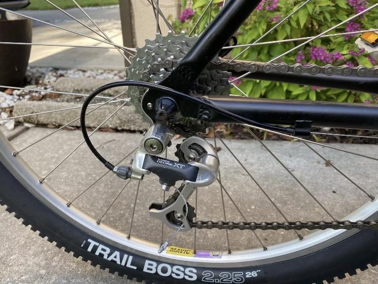 Specialized Specialized Stumpjumper A1 FS Mountain Bike: Close-up view of a bicycle's rear drivetrain, featuring the cassette, derailleur, and chain on a black bike frame. The background includes a concrete surface and some greenery with purple flowers. The tire is labeled "TRAIL BOSS 2.25 26”.