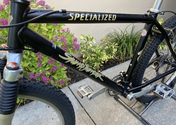 Specialized Specialized Stumpjumper A1 FS Mountain Bike: A close-up view of a black Specialized mountain bike featuring the "Stumpjumper" model name in gold lettering. The bike is positioned on a concrete surface surrounded by greenery and purple flowers, showcasing its suspension fork and pedal design.