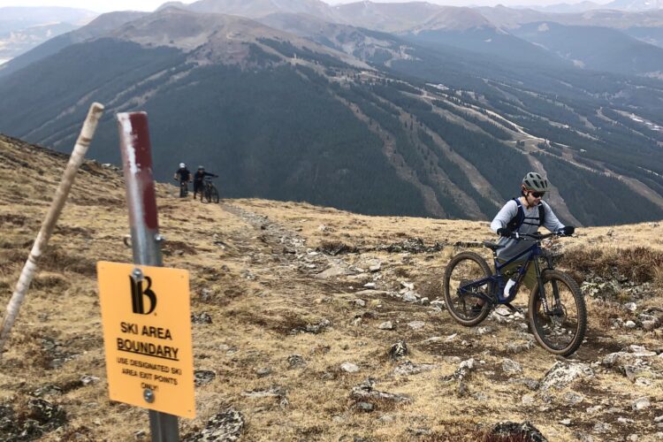 A mountain biker navigates a rocky trail near a ski area boundary sign, with a scenic mountainous landscape in the background. Two other bikers are visible further up the trail, surrounded by alpine terrain and trees.