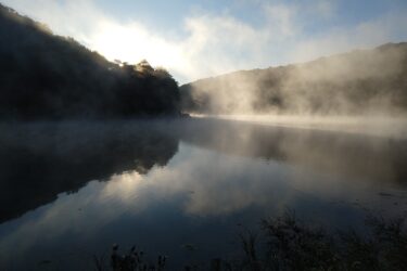 A serene landscape featuring a misty lake at sunrise, surrounded by hills. The water reflects the soft light and fog, creating a tranquil atmosphere with lush foliage in the foreground. Sidie Hollow mountain bike trail.