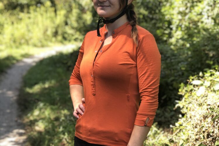 A woman wearing a black helmet and an orange long-sleeve shirt stands on a trail surrounded by greenery. She has her hands on her hips and is looking to the side, suggesting a relaxed yet ready posture for outdoor activities, likely cycling or mountain biking.