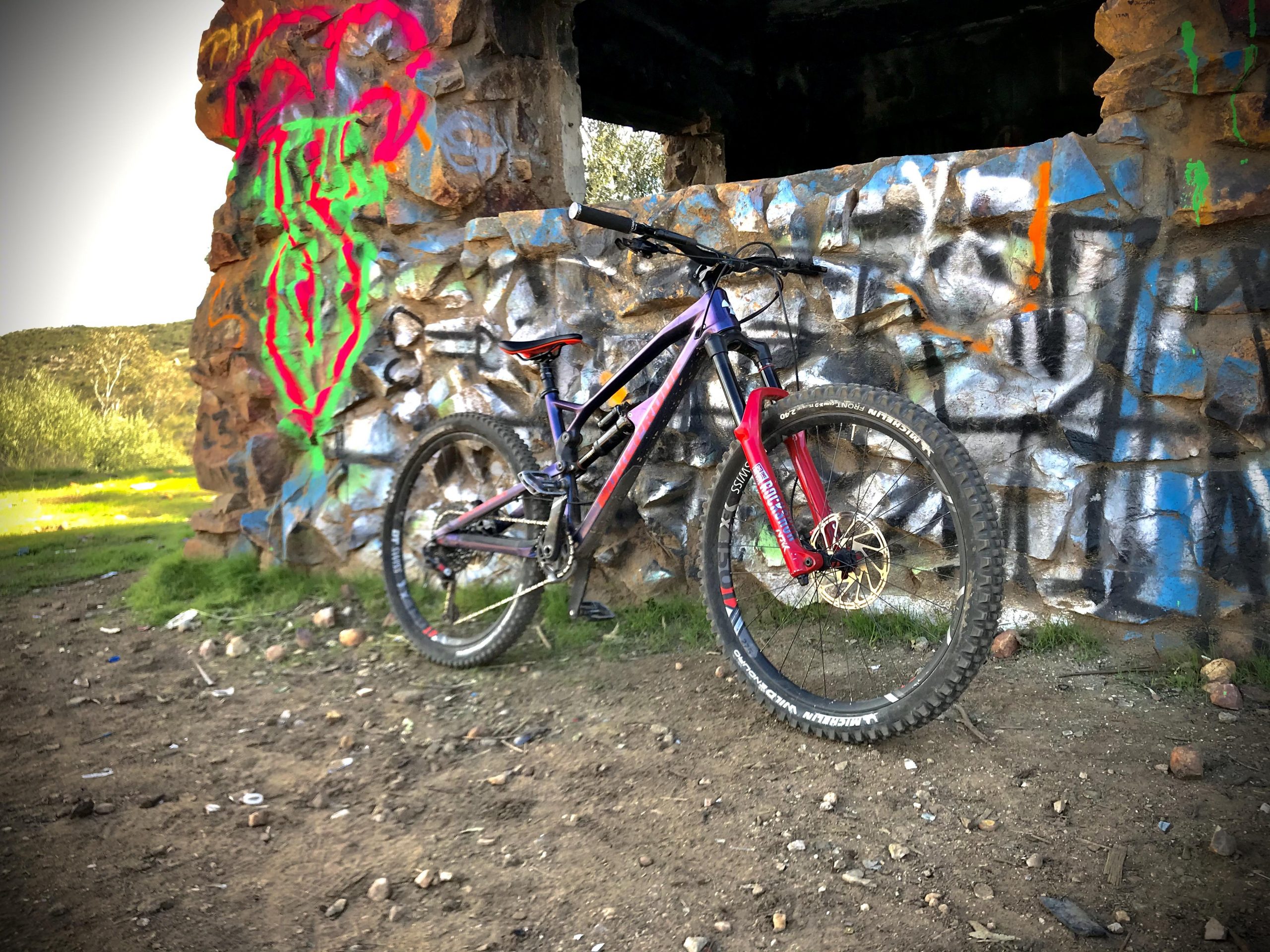 Nukeproof Mega AM 275 Pro: A mountain bike leaning against a graffiti-covered stone wall in a natural setting. The wall features colorful artwork in shades of green, pink, and blue, set against a backdrop of greenery and dirt ground.