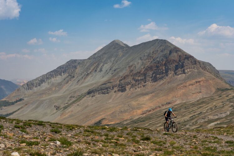 A mountain biker riding along a rugged, rocky landscape with towering mountains in the background under a clear blue sky. The scene captures the vastness of the terrain and the athlete's determination as they navigate the steep slopes.