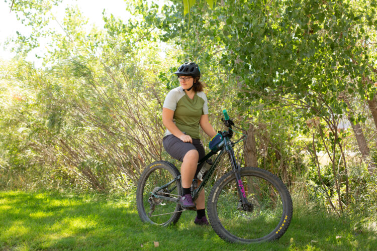 A person wearing a helmet and casual biking attire is sitting on a mountain bike in a grassy area surrounded by trees and foliage. The individual appears relaxed, looking off to the side. Sunlight filters through the leaves, creating a natural outdoor setting.