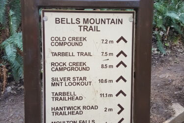Signpost for the Bells Mountain Trail, listing distances to various points including Cold Creek Compound (7.2 km), Tarbell Trail (7.5 km), Rock Creek Campground (8.5 km), Silver Star Mountain Lookout (10.6 km), Tarbell Trailhead (11.1 km), Hantwick Road Trailhead (2 km), and Moulton Falls Park Bridge (0.4 km). The background features lush green ferns and trees. Bell Mountain Trail mountain bike trail.