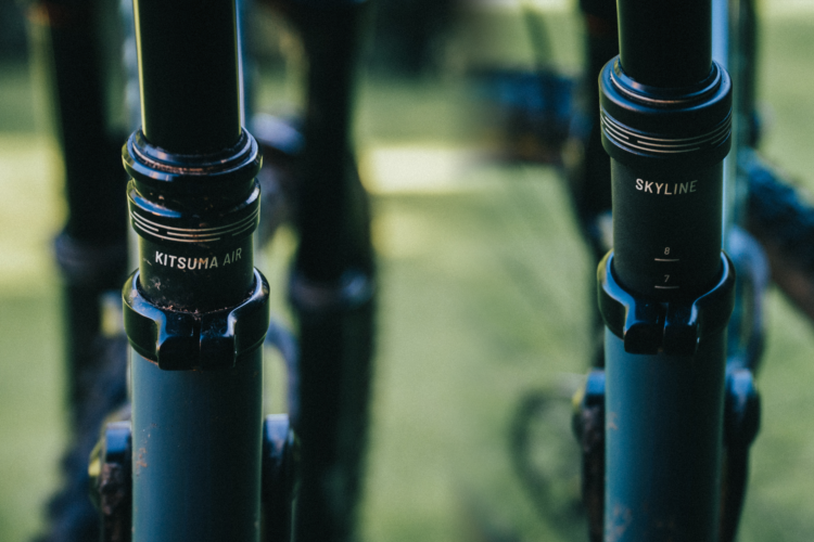 Close-up of two bicycle suspension forks labeled "KITSUMA AIR" and "SKYLINE," with visible adjustment markings and a blurred green background.