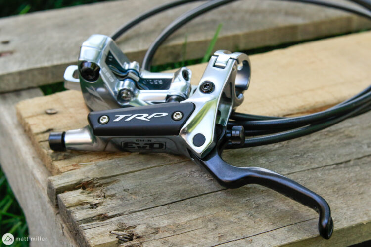 A close-up view of a TRP bicycle brake lever and caliper, featuring a sleek metallic finish and black accents, resting on weathered wooden planks. The image showcases the brake line and lever design, typically used for mountain or downhill biking.