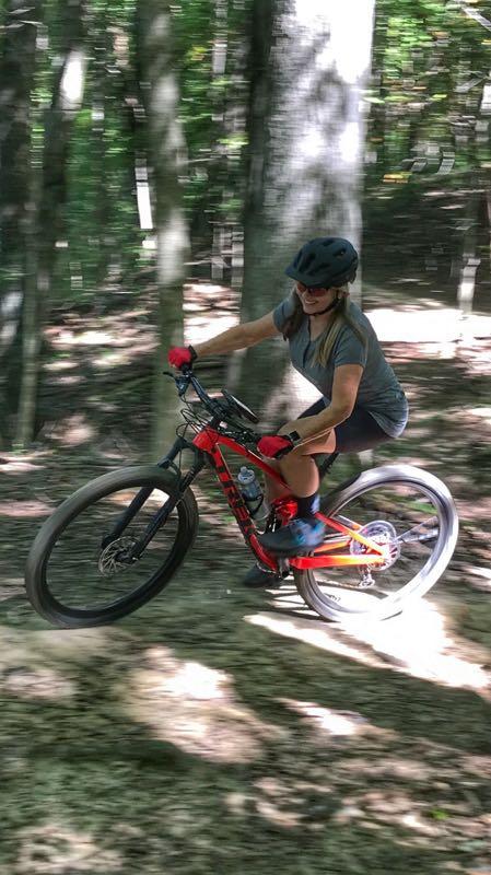 A person riding a mountain bike on a trail through a wooded area, captured mid-motion with a focus on speed and agility. The rider is wearing a helmet and has a determined expression, showcasing a sense of adventure in a natural, lush environment. East Fork mountain bike trail.