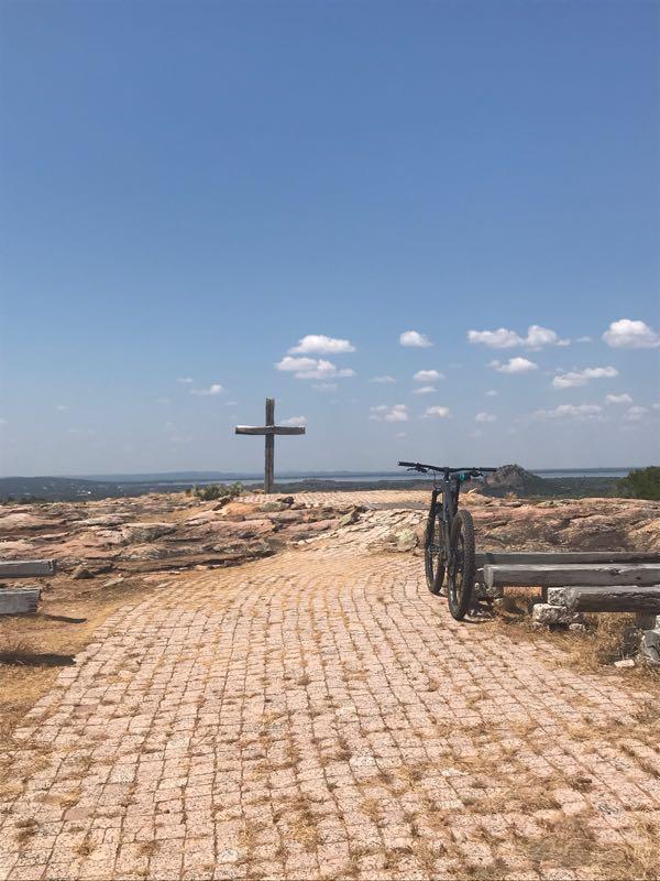 A mountain bike parked on a paved path leading to a wooden cross atop a rocky outcrop, with a panoramic view of the landscape under a clear blue sky with scattered clouds. Reveille Peak Ranch mountain bike trail.