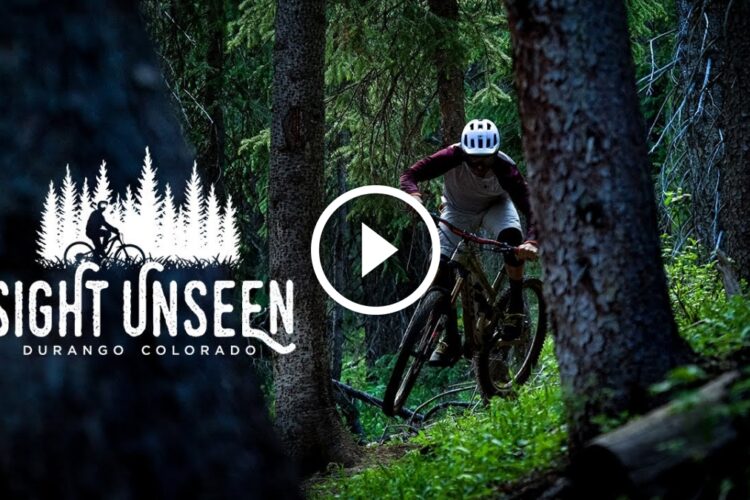 A mountain biker navigating through a dense forest trail, surrounded by tall pine trees, with the text "Sight Unseen Durango Colorado" and a play button icon overlayed on the image. The lighting is dim, emphasizing the lush greenery and the dynamic action of the biker.