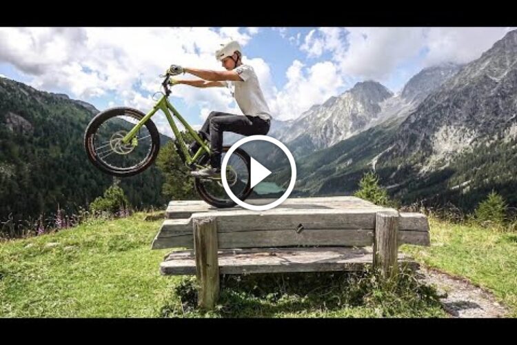 A person performs a bike stunt on a wooden bench, lifting the front wheel off the ground, with a scenic mountain landscape in the background. The sky is partly cloudy, and greenery surrounds the area.