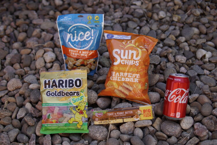 A variety of snacks arranged on a bed of pebbles, including a bag of Nice! Fruit and Nut Trail Mix, a package of Haribo Sour Goldbears, a bag of Sun Chips in Harvest Cheddar flavor, a Keebler Toast and Peanut Butter snack pack, and a can of Coca-Cola.