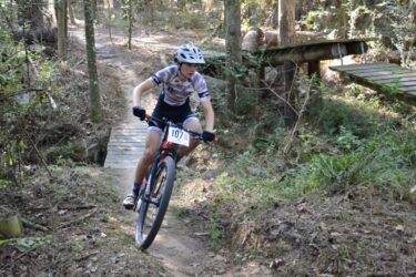 A young cyclist in a racing uniform navigates a narrow dirt trail alongside a wooden bridge in a forested area. The cyclist is focused and leaning forward on the bike, with a race number displayed on the front. Surrounding vegetation includes trees and underbrush. Mt. Zion Bike Trails mountain bike trail.