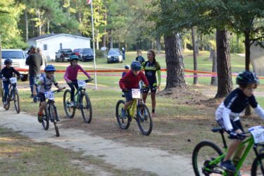 A group of children riding bicycles on a dirt path during a cycling event, surrounded by trees and spectators. The riders are wearing helmets and race numbers, and there are flags and vehicles in the background. Mt. Zion Bike Trails mountain bike trail.