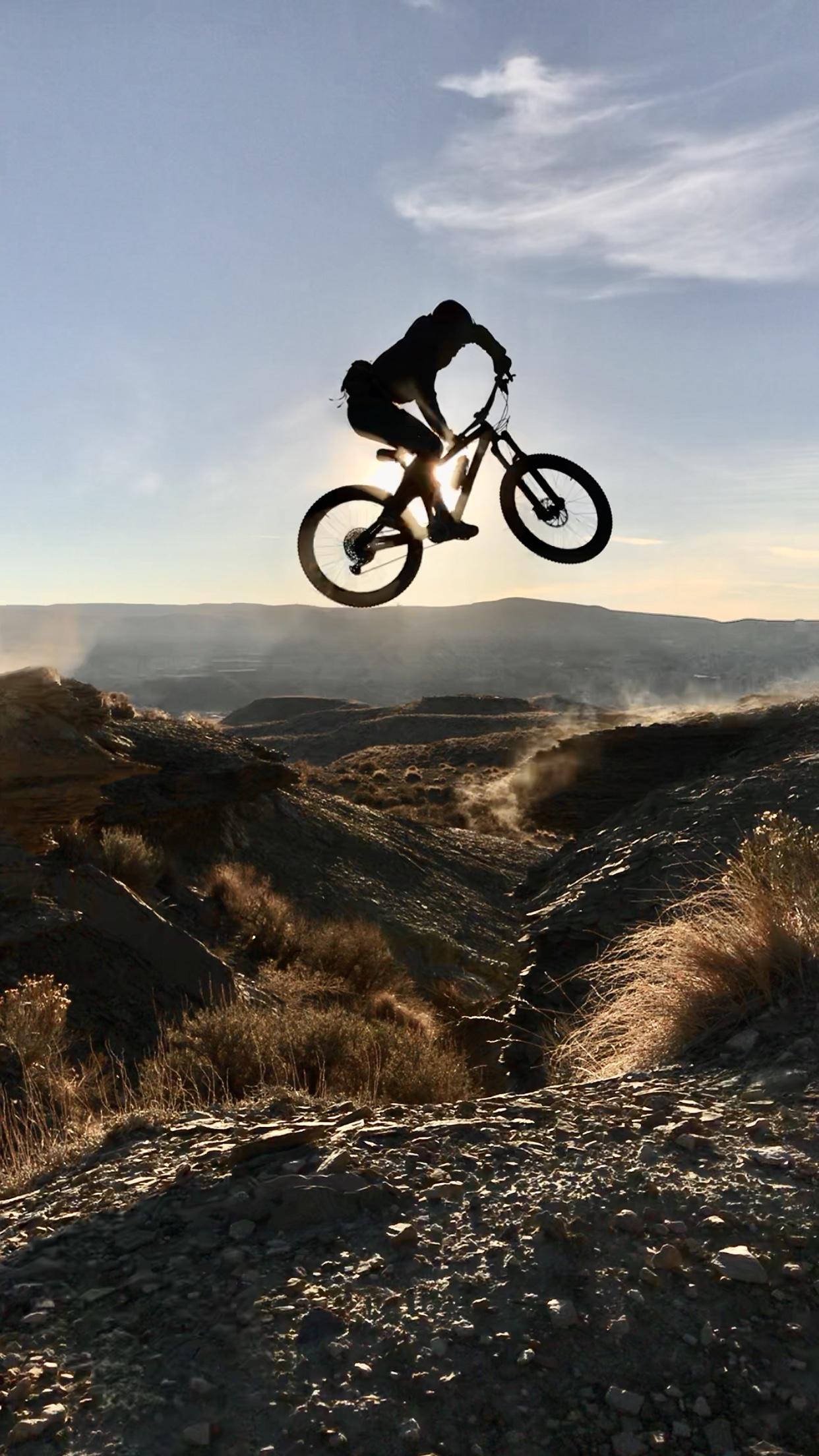 A silhouette of a mountain biker jumping over a rocky terrain against a backdrop of a sunset sky, with distant hills and smoke rising from the ground. Wilkins Peak Trails mountain bike trail.