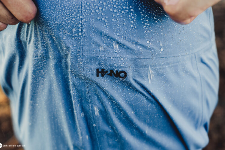 A close-up image of a person's shirt with water droplets clearly visible on the fabric, showcasing the material's water-resistant properties. The shirt is light blue with a logo featuring the text "H2No" displayed prominently. The person's hand is partially visible, holding the fabric.