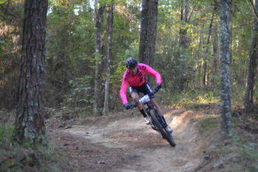 A mountain biker in a bright pink long-sleeve jersey and black shorts leans into a turn on a dirt trail surrounded by trees in a forested area. The cyclist is wearing a helmet and is focused on navigating the path. Mt. Zion Bike Trails mountain bike trail.