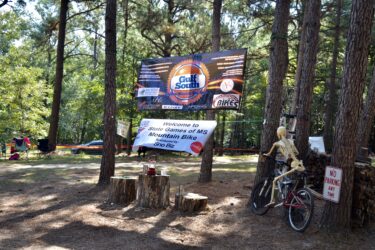Banner display for the State Games of Mississippi Mountain Bike event, featuring sponsors' logos, a skeleton posed on a bicycle, and wooden stumps serving as seating. Surrounded by trees in a natural setting, with additional seating visible in the background. Mt. Zion Bike Trails mountain bike trail.