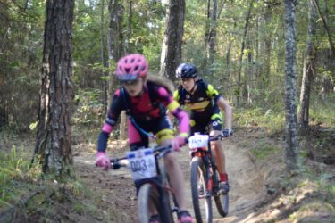 Two young cyclists navigate a dirt trail in a forest setting. One rider wears a pink helmet and a black and red cycling outfit, while the other is in a black and yellow cycling jersey. Surrounding them are trees and lush greenery, suggesting an outdoor race or biking event. Mt. Zion Bike Trails mountain bike trail.