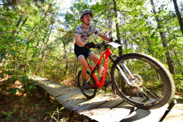 A young cyclist riding a red mountain bike on a wooden bridge through a lush, green forest. The cyclist is focused and wearing a helmet, showcasing determination as they navigate the trail. Sunlight filters through the trees, creating a vibrant and energetic atmosphere. Mt. Zion Bike Trails mountain bike trail.