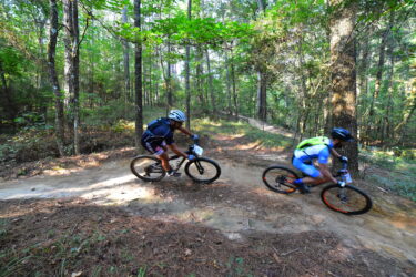 Two mountain bikers navigating a winding trail in a forested area. The riders, wearing helmets and cycling gear, are captured in motion as they maneuver around trees and foliage, highlighting an active outdoor environment. Mt. Zion Bike Trails mountain bike trail.