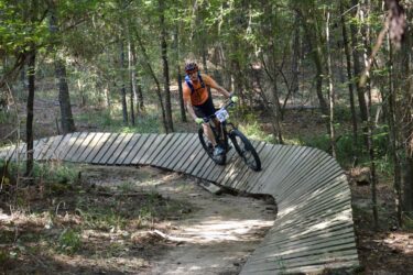 A mountain biker riding on a wooden trail through a forest, navigating a curved section of the path. The cyclist is wearing an orange shirt, a helmet, and gloves, with a race number visible on their bike. Surrounding trees and underbrush provide a natural backdrop. Mt. Zion Bike Trails mountain bike trail.