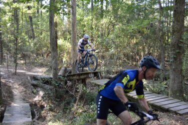 Two mountain bikers riding through a forested trail. One biker is navigating a narrow wooden bridge while the other is in motion on a nearby path, surrounded by trees and greenery. The scene captures the thrill of mountain biking in a natural setting. Mt. Zion Bike Trails mountain bike trail.
