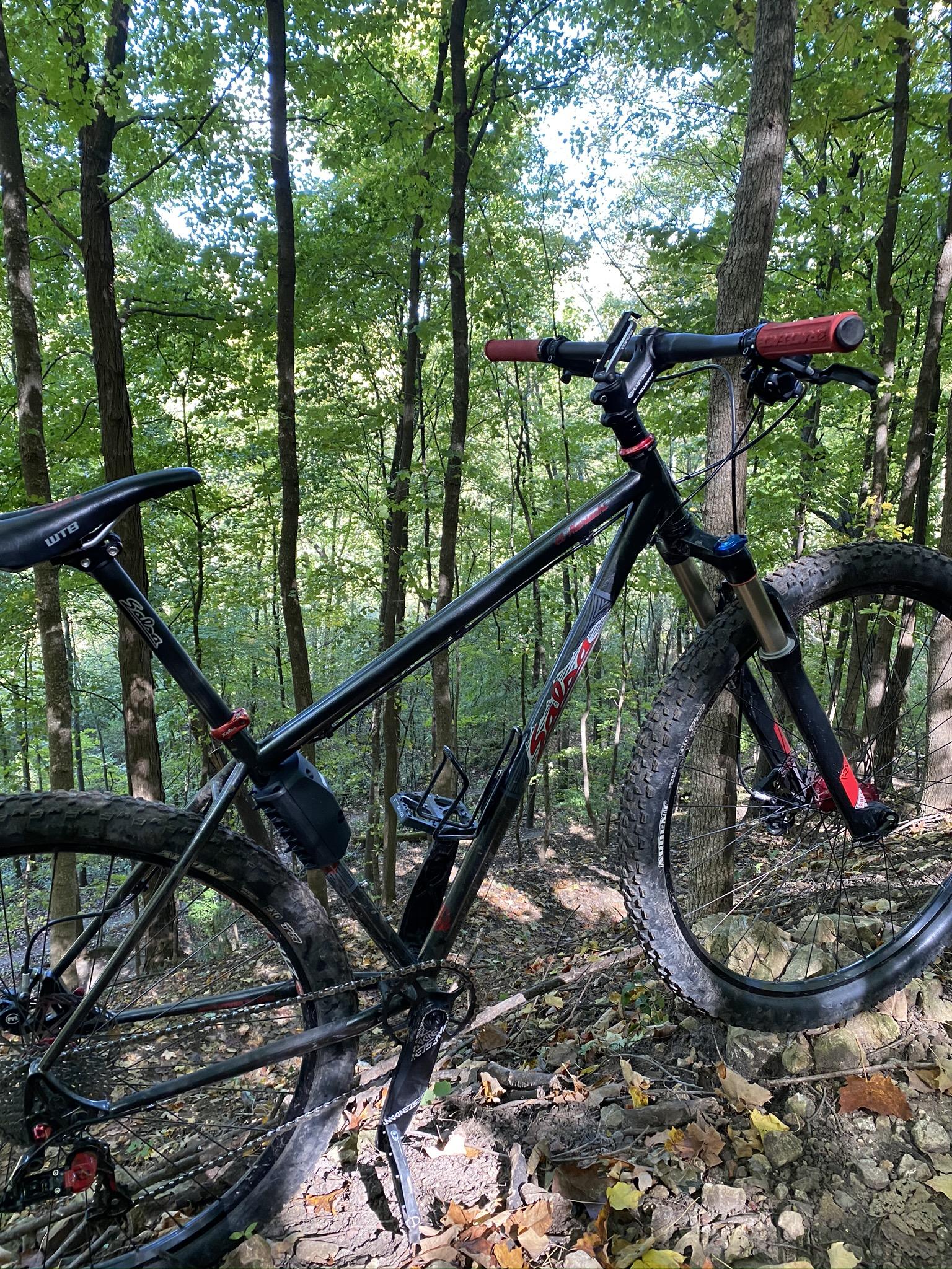 Salsa El Mariachi: A black mountain bike with red handles resting on a rocky trail surrounded by green trees and foliage. Sunlight filters through the leaves, creating a dappled light effect on the ground.