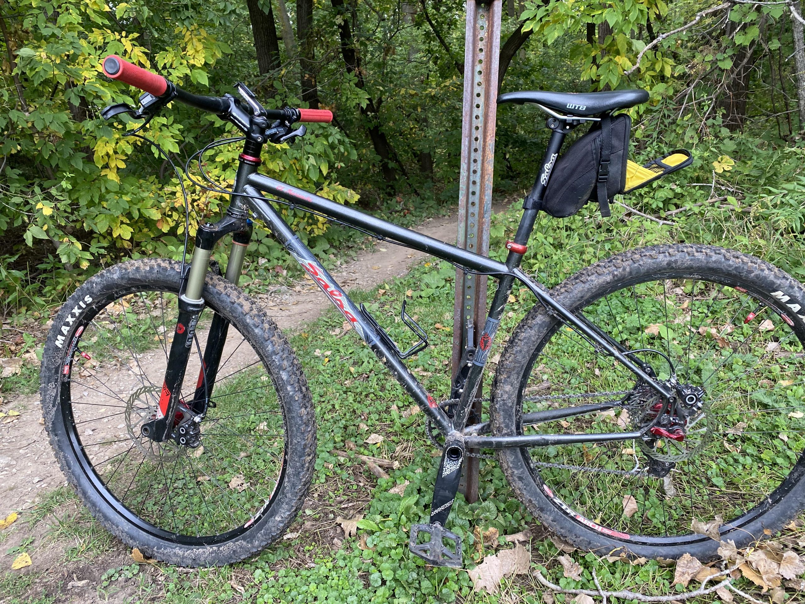 Salsa El Mariachi: A mountain bike with red handlebars and a black frame is parked next to a metal signpost surrounded by green foliage. The bike features thick, knobby tires and a small black saddlebag attached to its seat. A dirt path can be seen winding through the trees in the background.