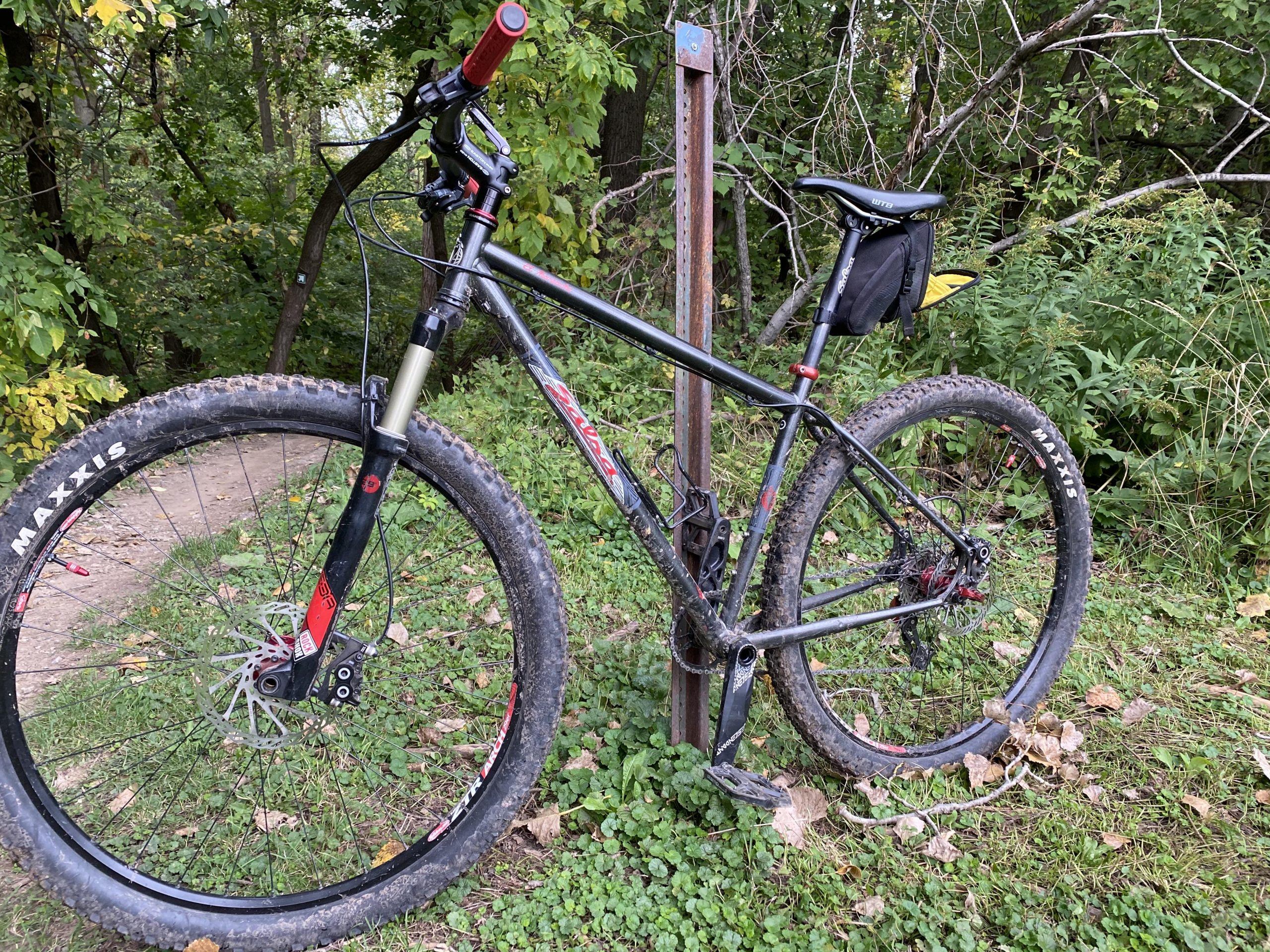 Salsa El Mariachi: A mountain bike with a sturdy frame and knobby tires, parked next to a trail sign in a wooded area. The bike is covered in mud, indicating recent off-road use. Lush greenery and trees surround the scene, suggesting a natural outdoor environment ideal for biking.