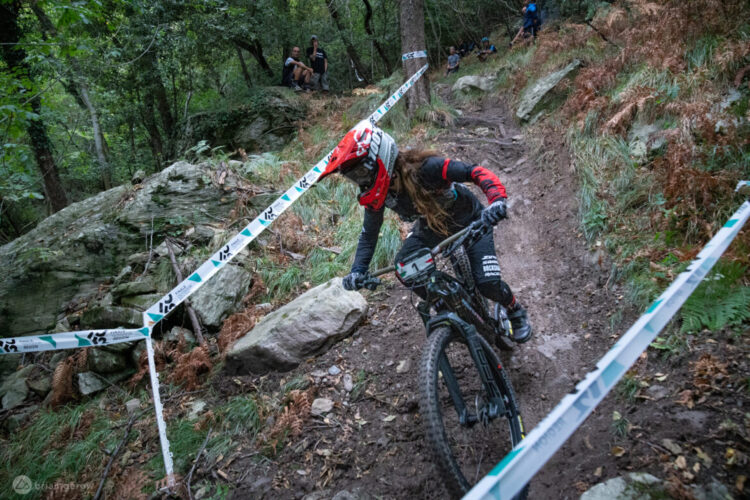 A mountain biker navigating a muddy, rocky trail in a forested area. The rider wears a red helmet and black gear, while a few spectators watch from the side. Course markers are visible along the trail, adding a competitive atmosphere.