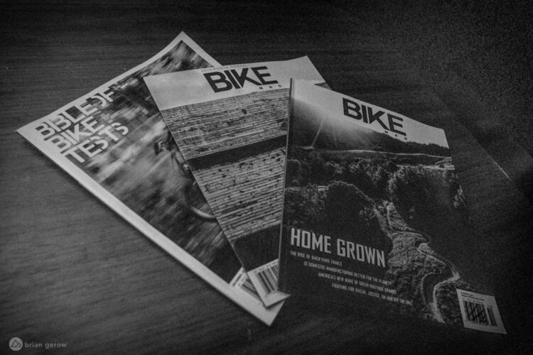Three issues of a bike magazine are stacked on a wooden surface. The magazines feature bold titles such as "BIKE" and "Bible of Bike Tests," along with a cover article titled "Home Grown." The overall image is in black and white, highlighting the textures and details of the magazines and the grain of the wood beneath them.