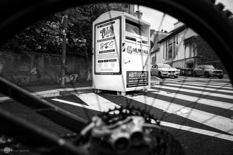 A black and white photograph depicting a clothing donation bin labeled "Humana" situated on a street corner. The image is framed from a low angle, showing part of a bicycle wheel in the foreground and intersecting lines of a crosswalk. In the background, cars are visible along the street, alongside a wall covered in greenery.