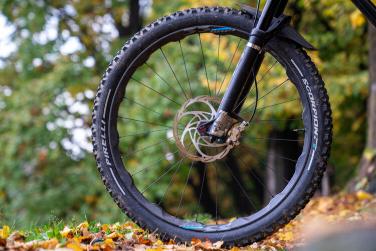 A close-up view of a mountain bike wheel, featuring a Pirelli tire with pronounced tread for off-road traction. The wheel is positioned in a natural setting with scattered autumn leaves on the ground, and blurred greenery in the background. The disc brake assembly is visible, showcasing its intricate design.