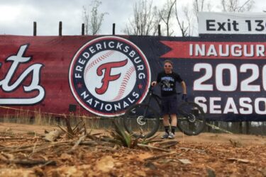 A young person stands next to a bicycle in front of a large banner for the Fredericksburg Nationals baseball team, advertising their inaugural 2020 season. The background features a cloudy sky and indications of a dirt area with sparse vegetation. Rappahanock River Trail mountain bike trail.